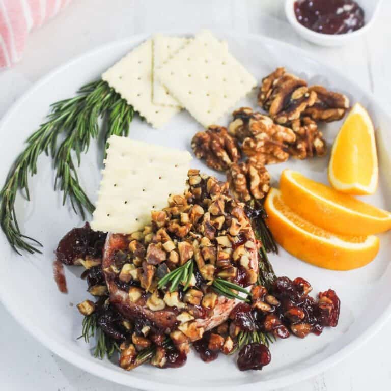 A white plate with crackers, walnuts, orange slices, sprigs of rosemary, and baked brie topped with chopped nuts and dried fruit. A small bowl of jam is in the background.