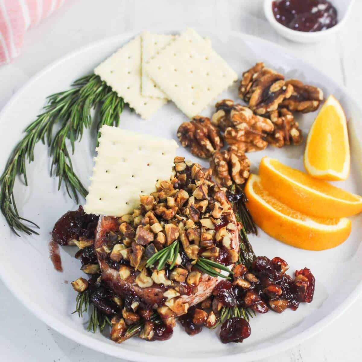 A white plate with crackers, walnuts, orange slices, sprigs of rosemary, and baked brie topped with chopped nuts and dried fruit. A small bowl of jam is in the background.