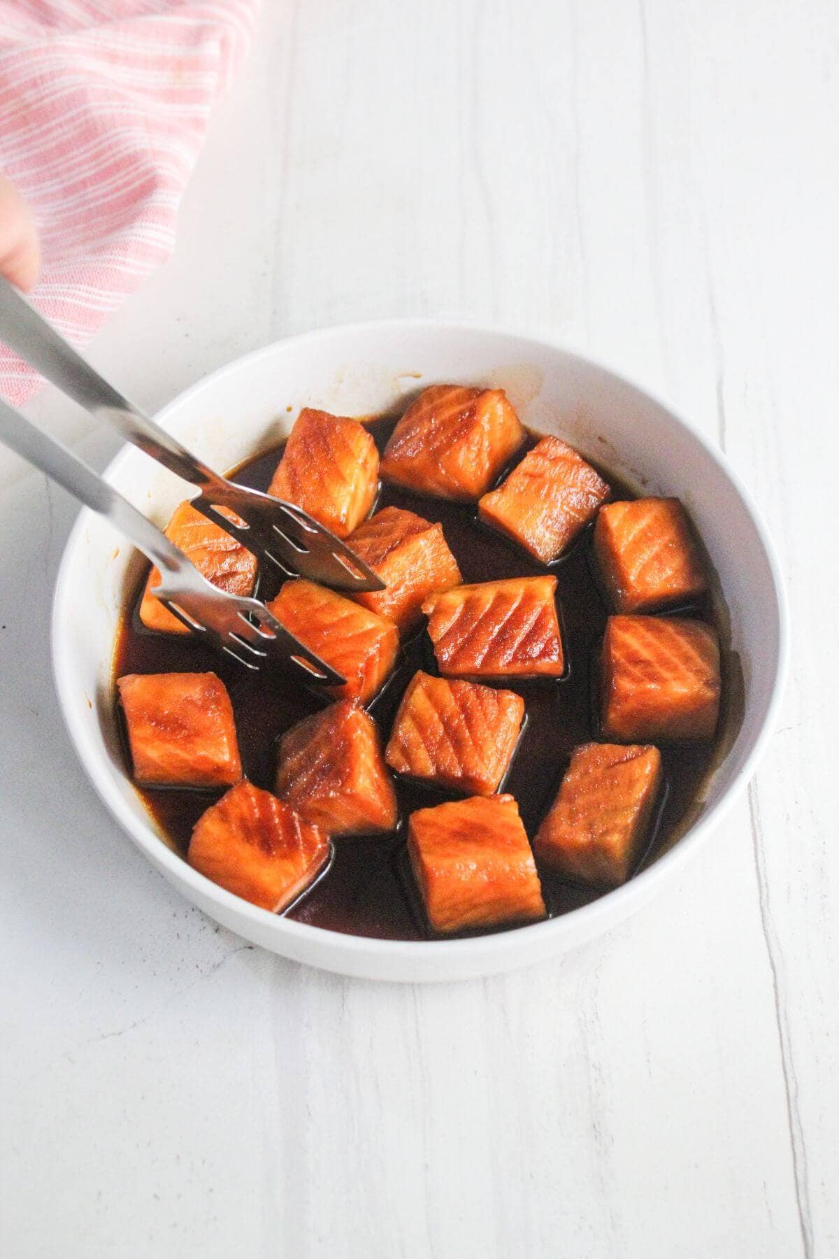 A white bowl filled with cubed salmon pieces in a dark sauce, with a fork and tongs picking up a piece.