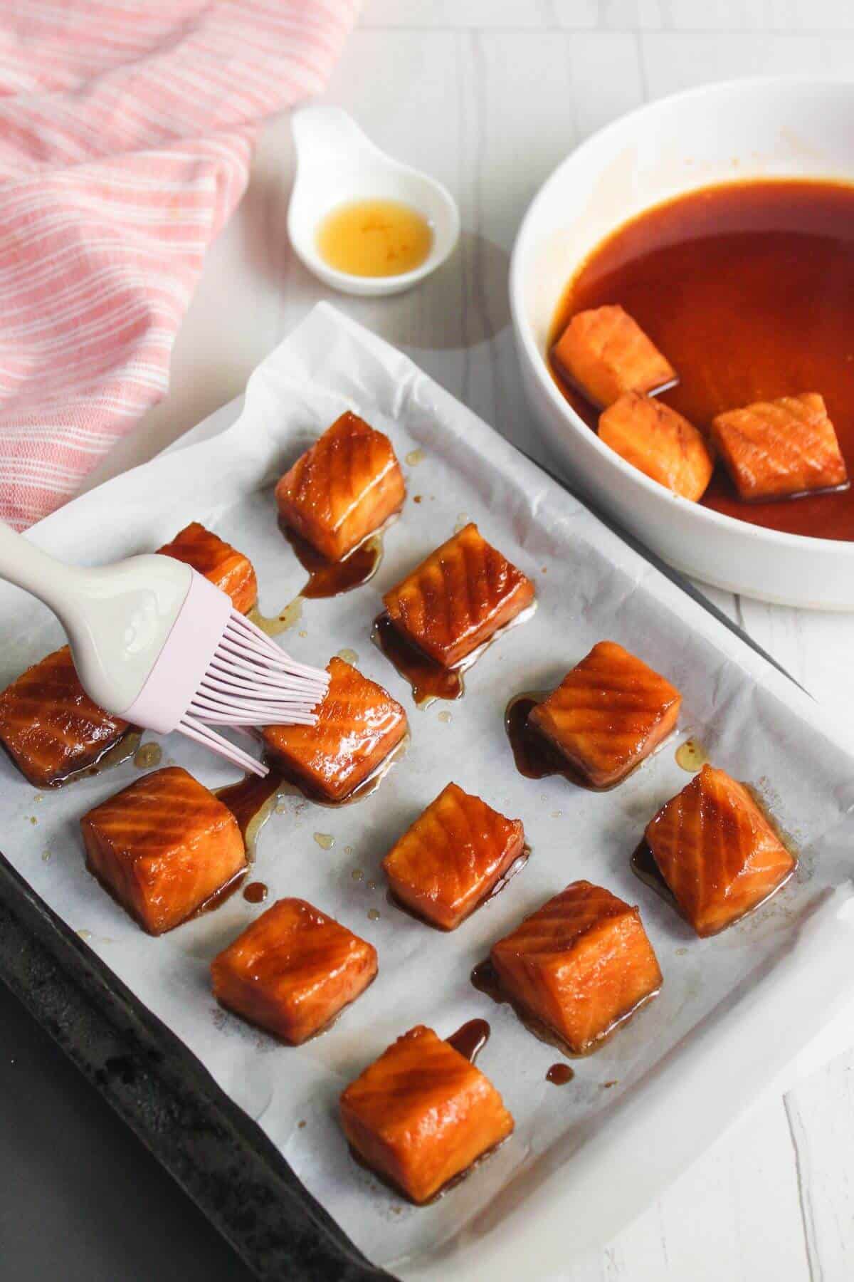 A baking tray lined with parchment holds cubed salmon pieces being brushed with a brown sauce. Nearby are a bowl with sauce and extra salmon, a spoon with liquid, and a pink cloth.