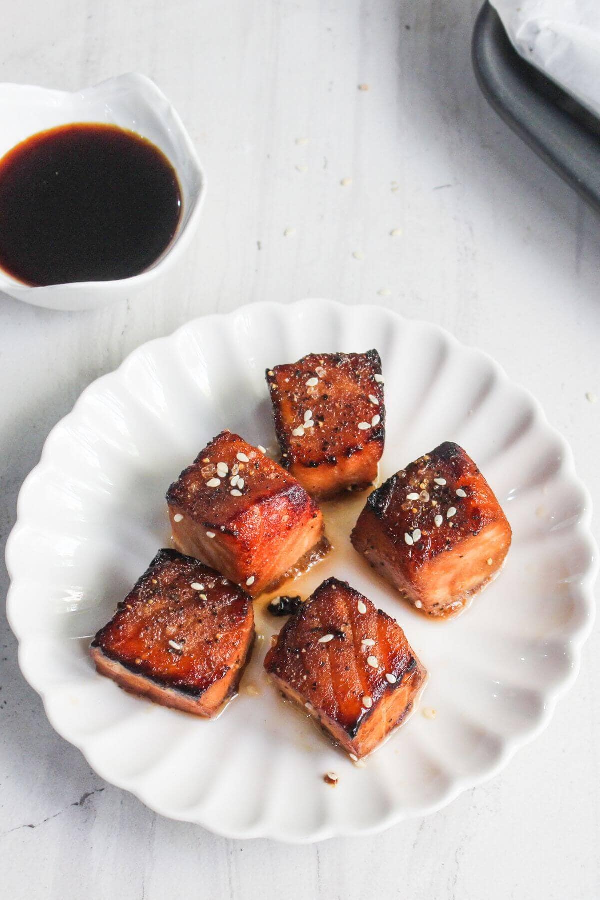 Five glazed and seared salmon cubes garnished with sesame seeds on a white scalloped plate, with a small bowl of dark dipping sauce in the background.