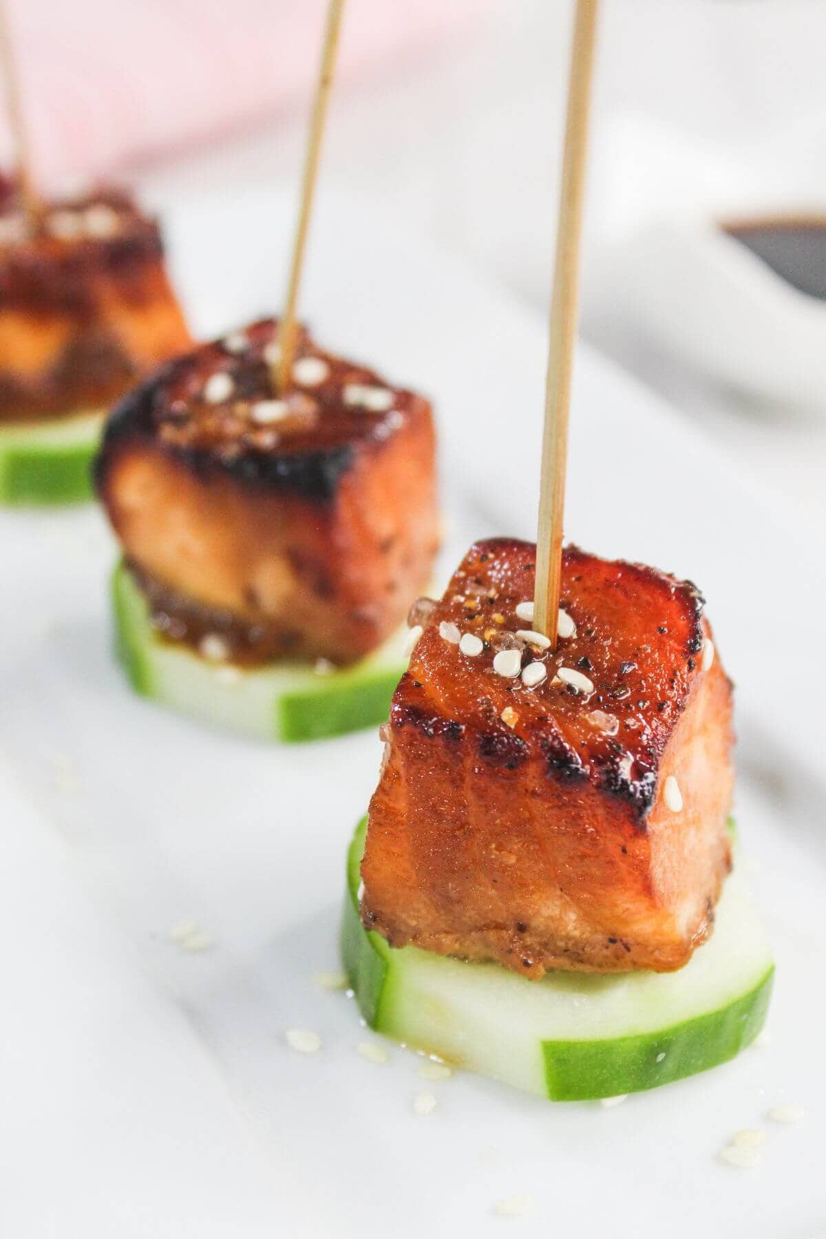Three skewered cubes of grilled tofu sit on cucumber slices, garnished with sesame seeds, arranged on a white surface.