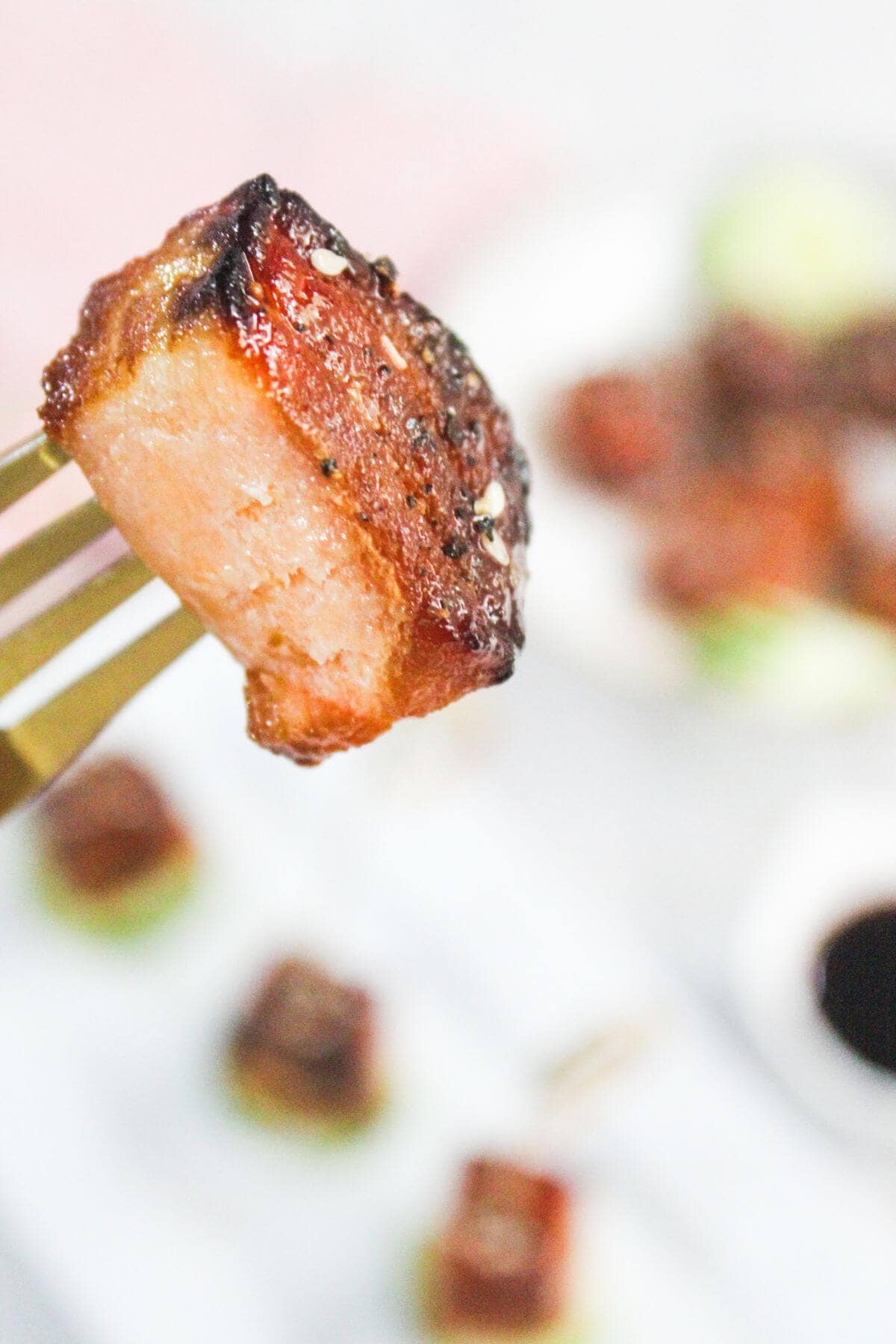 A close-up of a fork holding a glazed, seared piece of meat, with blurred food and sauce in the background.