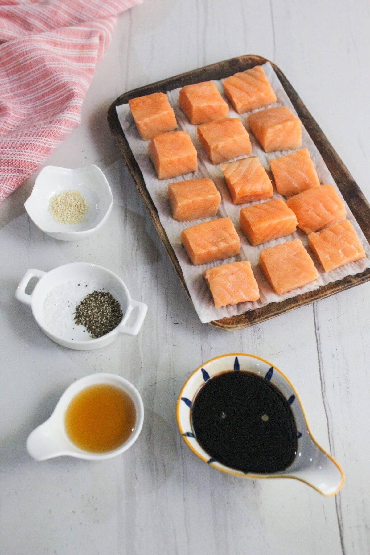 Cubed raw salmon on a tray with small bowls of sesame seeds, salt, pepper, oil, and soy sauce arranged on a white surface.