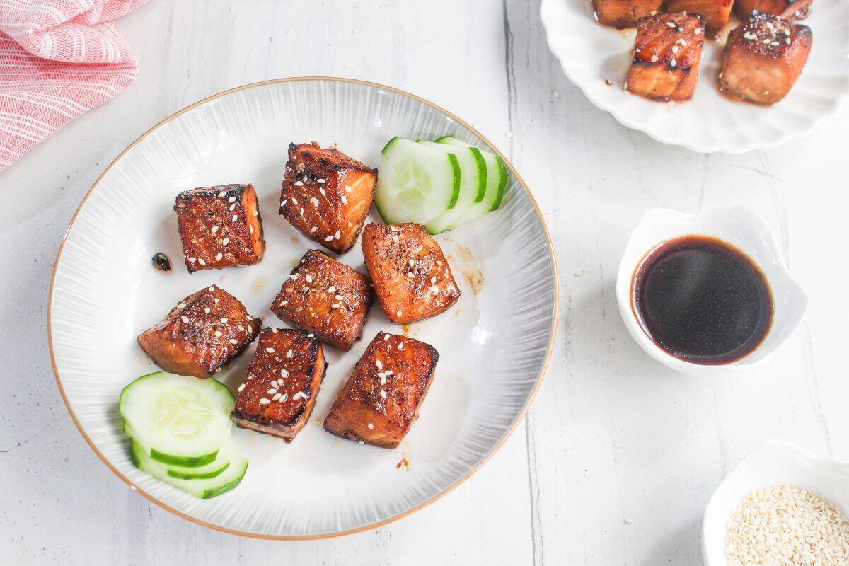 A plate of glazed salmon bites garnished with sesame seeds and cucumber slices, accompanied by a small bowl of soy sauce and a dish of sesame seeds.