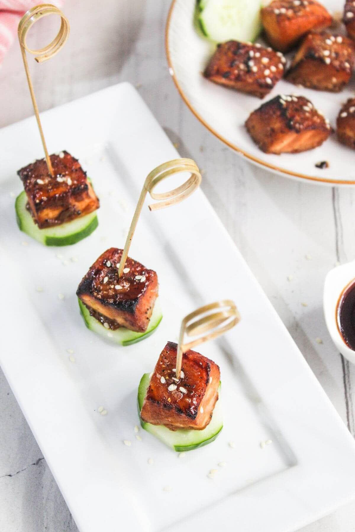 Three glazed tofu cubes on cucumber slices, each skewered with a decorative pick and sprinkled with sesame seeds, are arranged on a rectangular white plate.