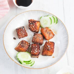 A plate with seven glazed salmon cubes garnished with sesame seeds, served with sliced cucumber on a white background.