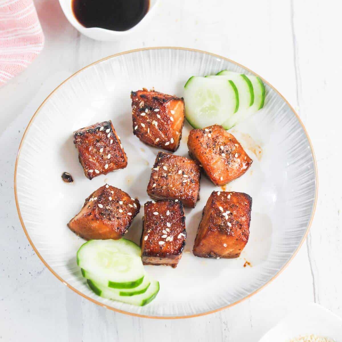 A plate with seven glazed salmon cubes garnished with sesame seeds, served with sliced cucumber on a white background.