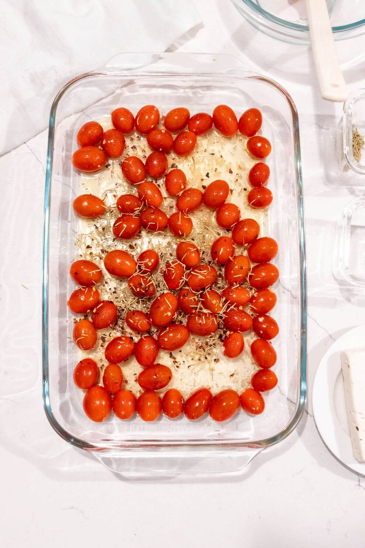 A glass baking dish with cherry tomatoes, garlic, and herbs arranged on top of a layer of uncooked pasta.