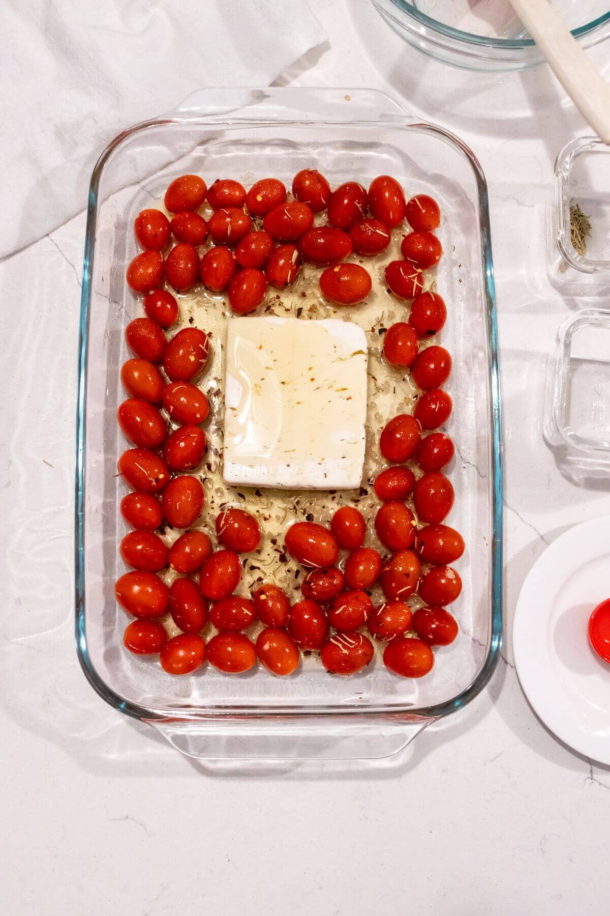 Glass baking dish with a block of feta cheese in the center, surrounded by cherry tomatoes and herbs, ready to be baked.