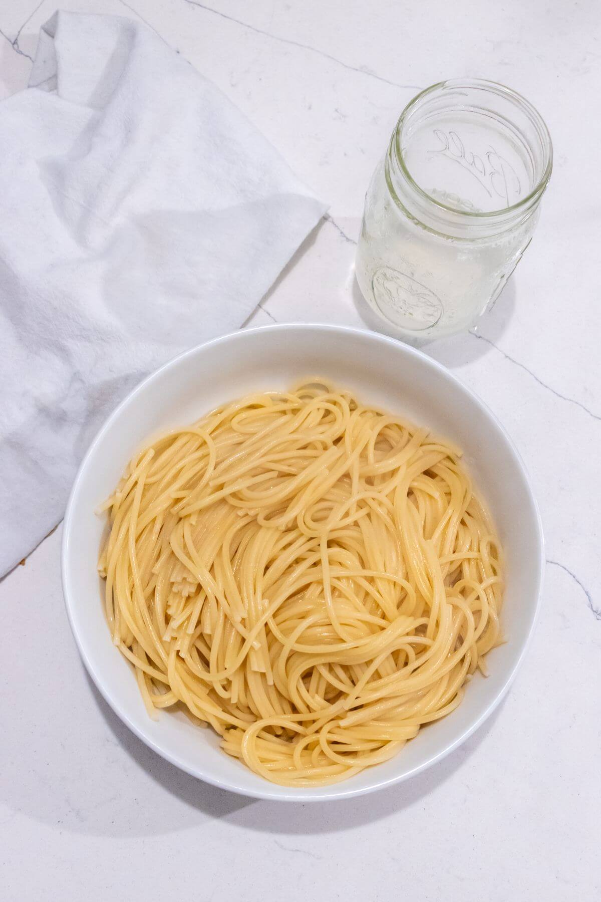 A white bowl of plain cooked spaghetti sits on a white surface next to a clear glass jar of water and a folded white cloth.