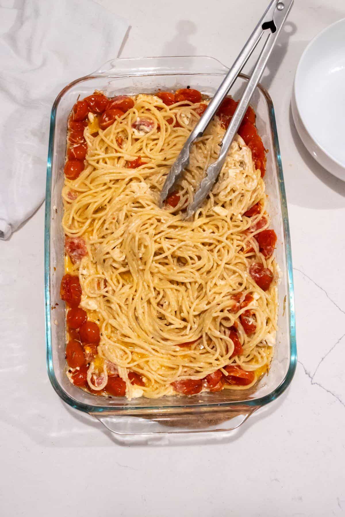 A glass baking dish containing baked spaghetti mixed with roasted cherry tomatoes and cheese, with metal tongs resting on top.