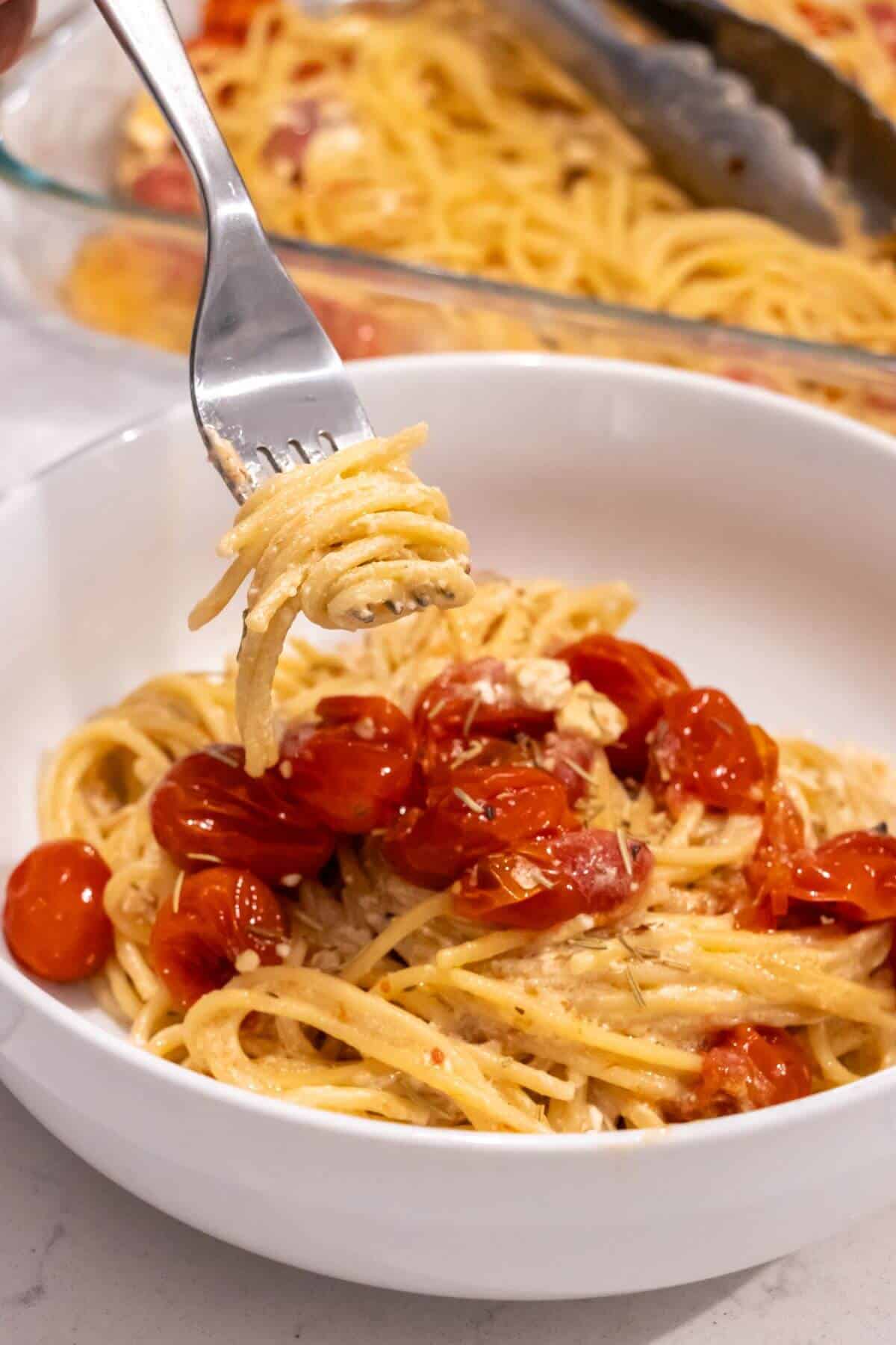 A white bowl of spaghetti topped with roasted cherry tomatoes and feta, with a fork twirling a bite of pasta above the bowl.
