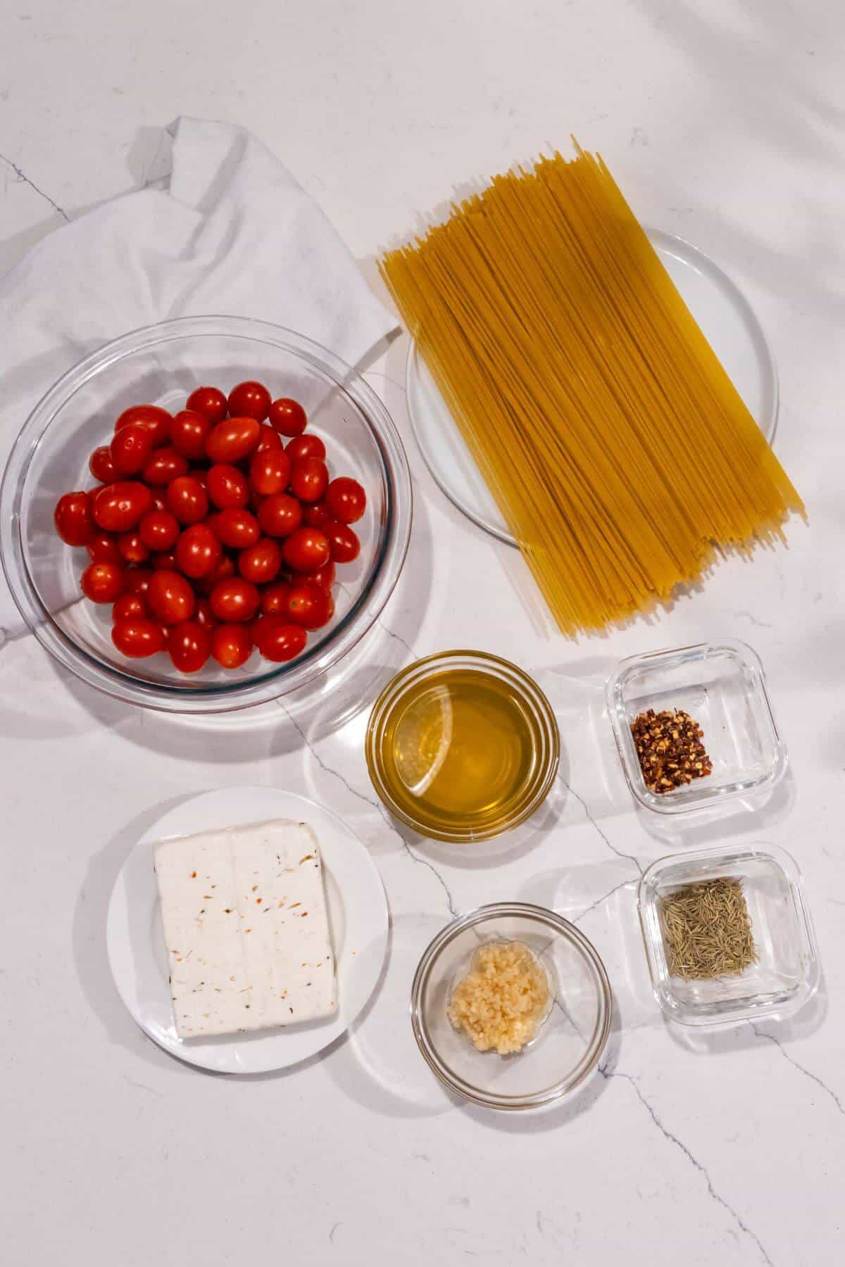 Uncooked spaghetti, cherry tomatoes, feta cheese, olive oil, minced garlic, dried rosemary, and red pepper flakes arranged in bowls on a white surface.