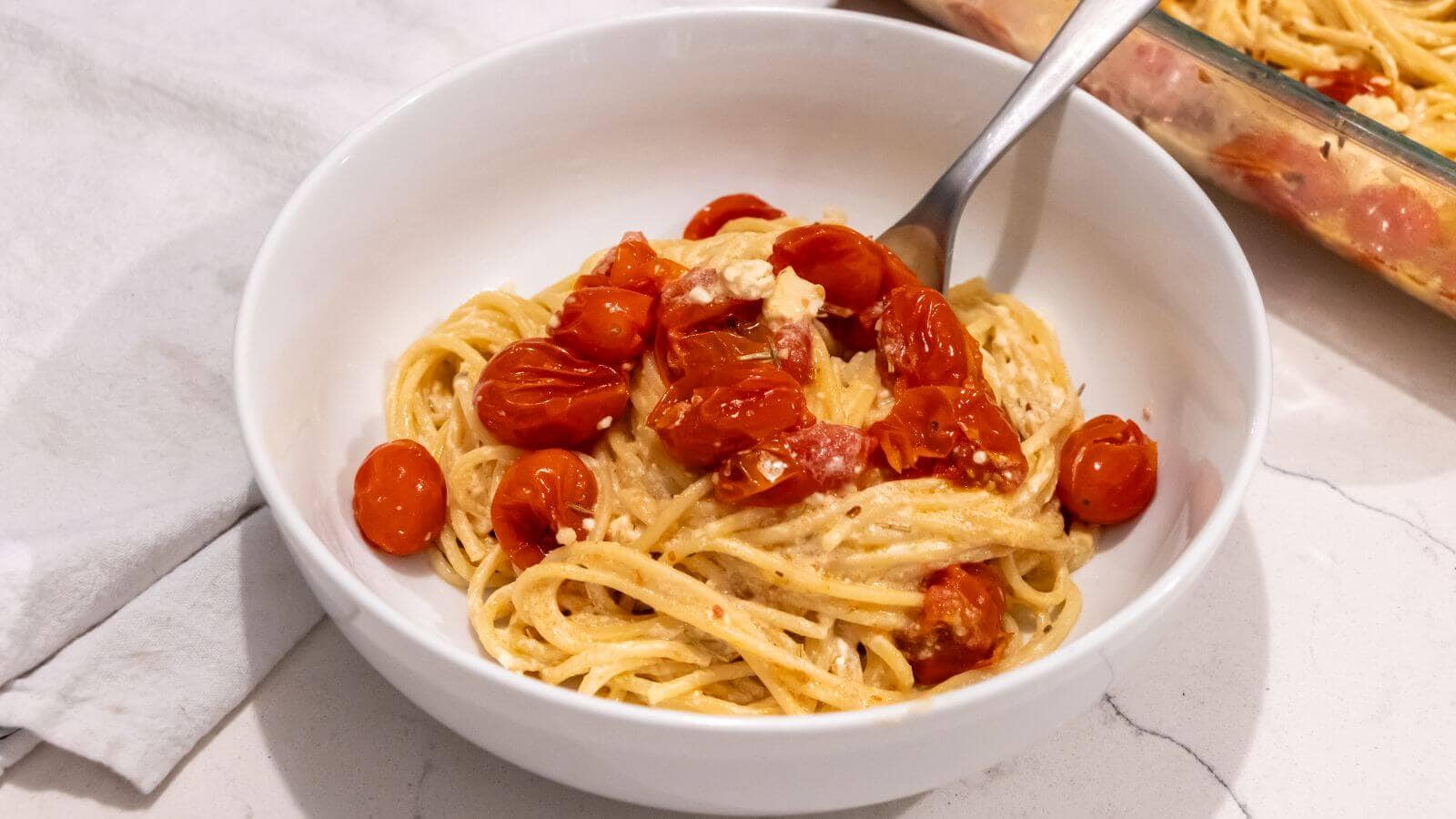 A white bowl filled with spaghetti tossed in a creamy sauce, topped with roasted cherry tomatoes and crumbled cheese, with a fork in the bowl.