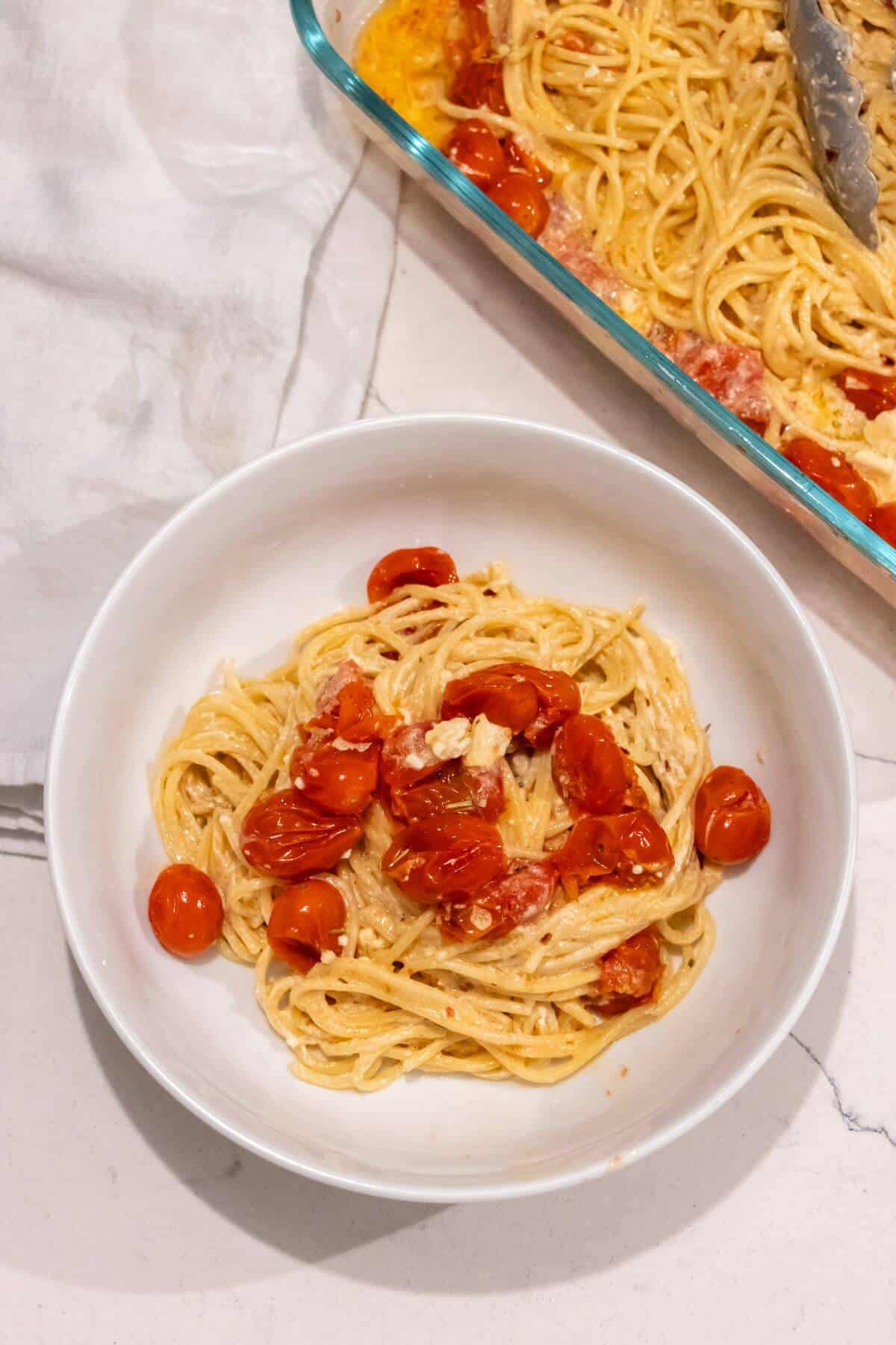 A bowl of spaghetti mixed with roasted cherry tomatoes and creamy sauce, with a baking dish of the same pasta in the background.