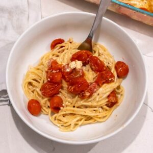 A bowl of spaghetti topped with roasted cherry tomatoes and crumbled cheese, with a fork placed in the bowl, on a marble countertop.