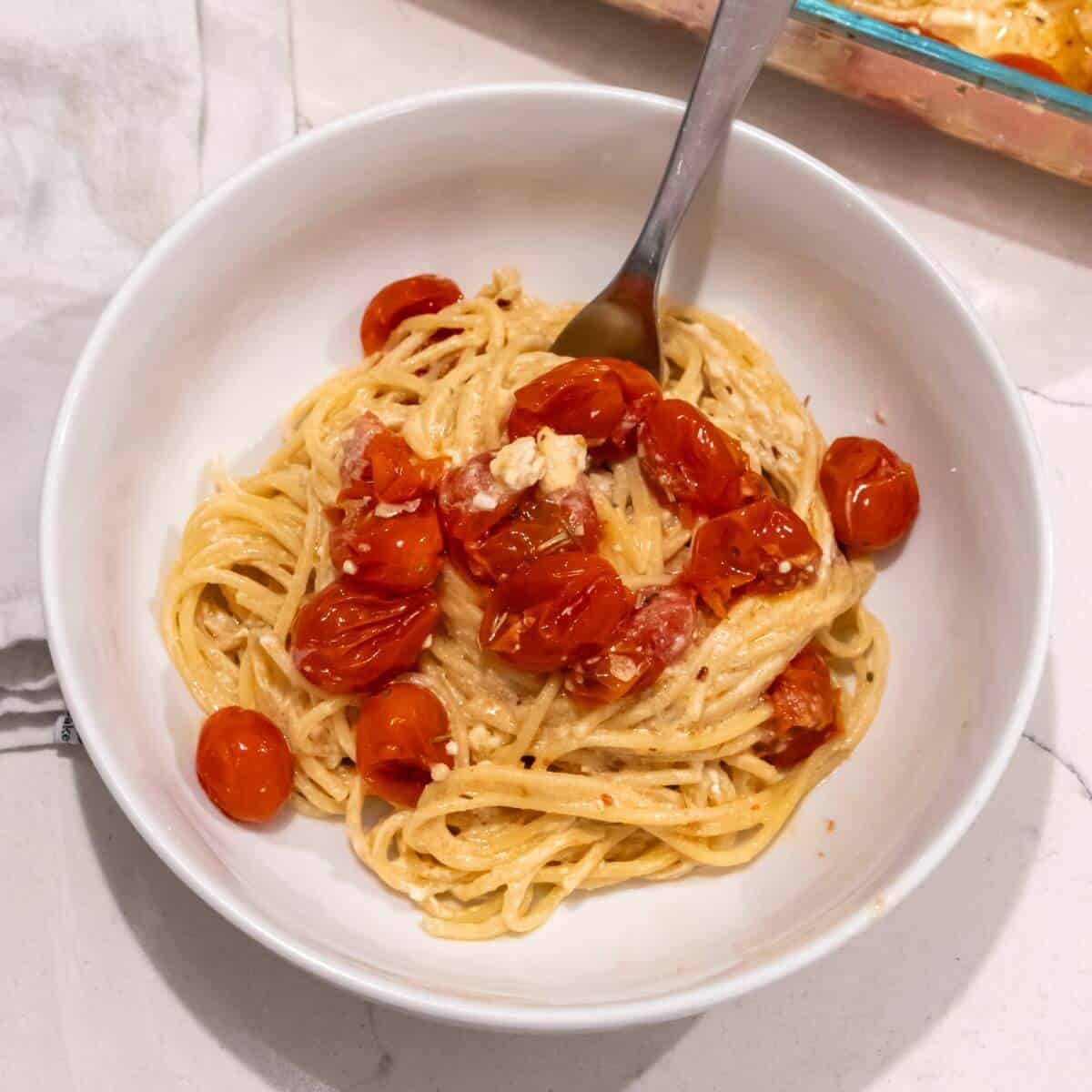 A bowl of spaghetti topped with roasted cherry tomatoes and crumbled cheese, with a fork placed in the bowl, on a marble countertop.