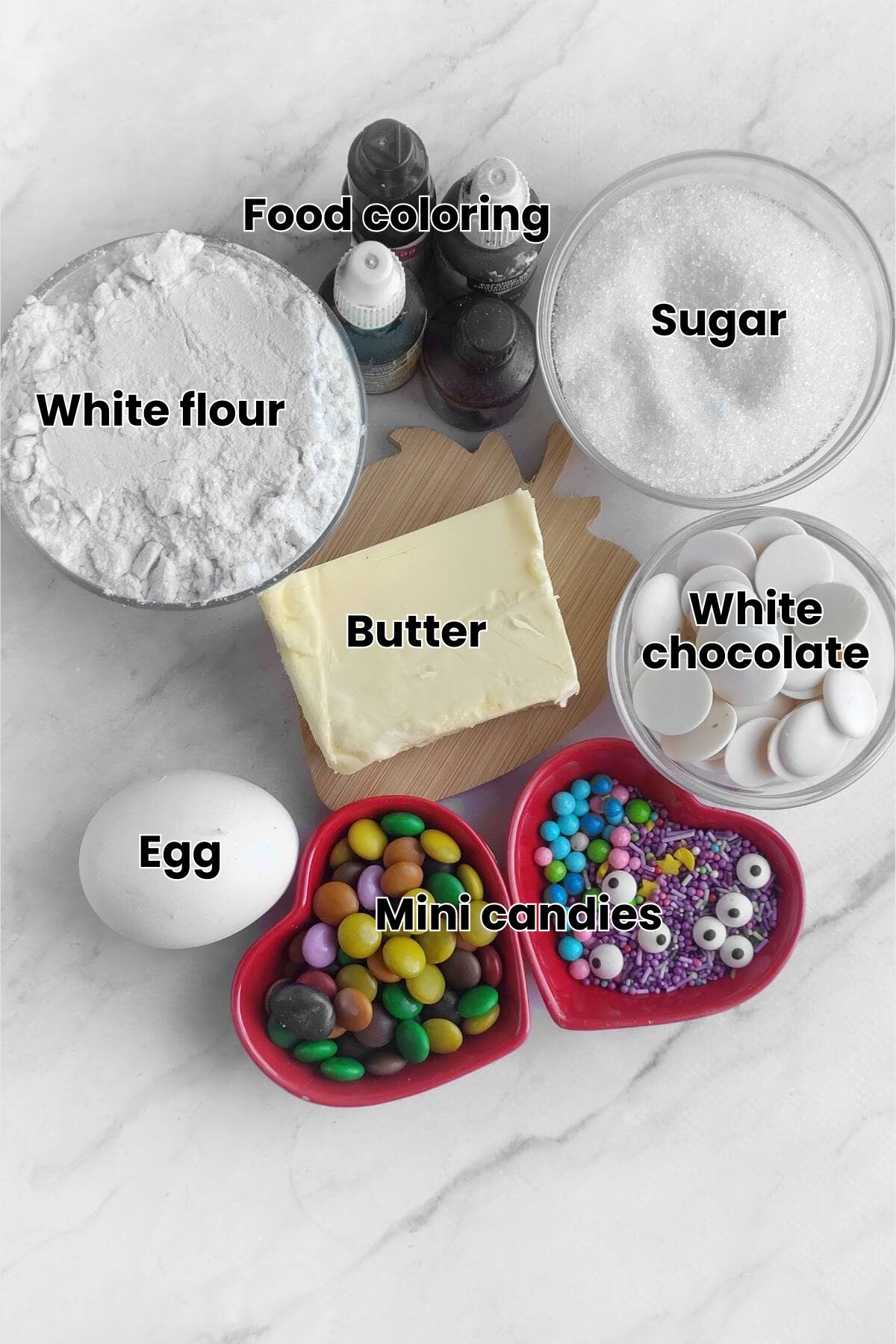 Baking ingredients on a counter including powdered sugar, food coloring, granulated sugar, butter, white candy melts, an egg, and two bowls of colorful candies and sprinkles.