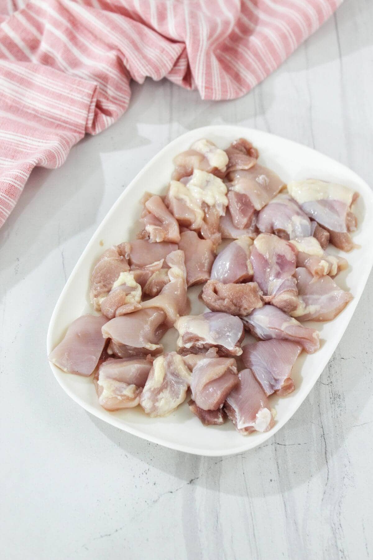 A white plate with raw, chopped pieces of chicken placed on a light-colored surface next to a pink and white striped cloth.