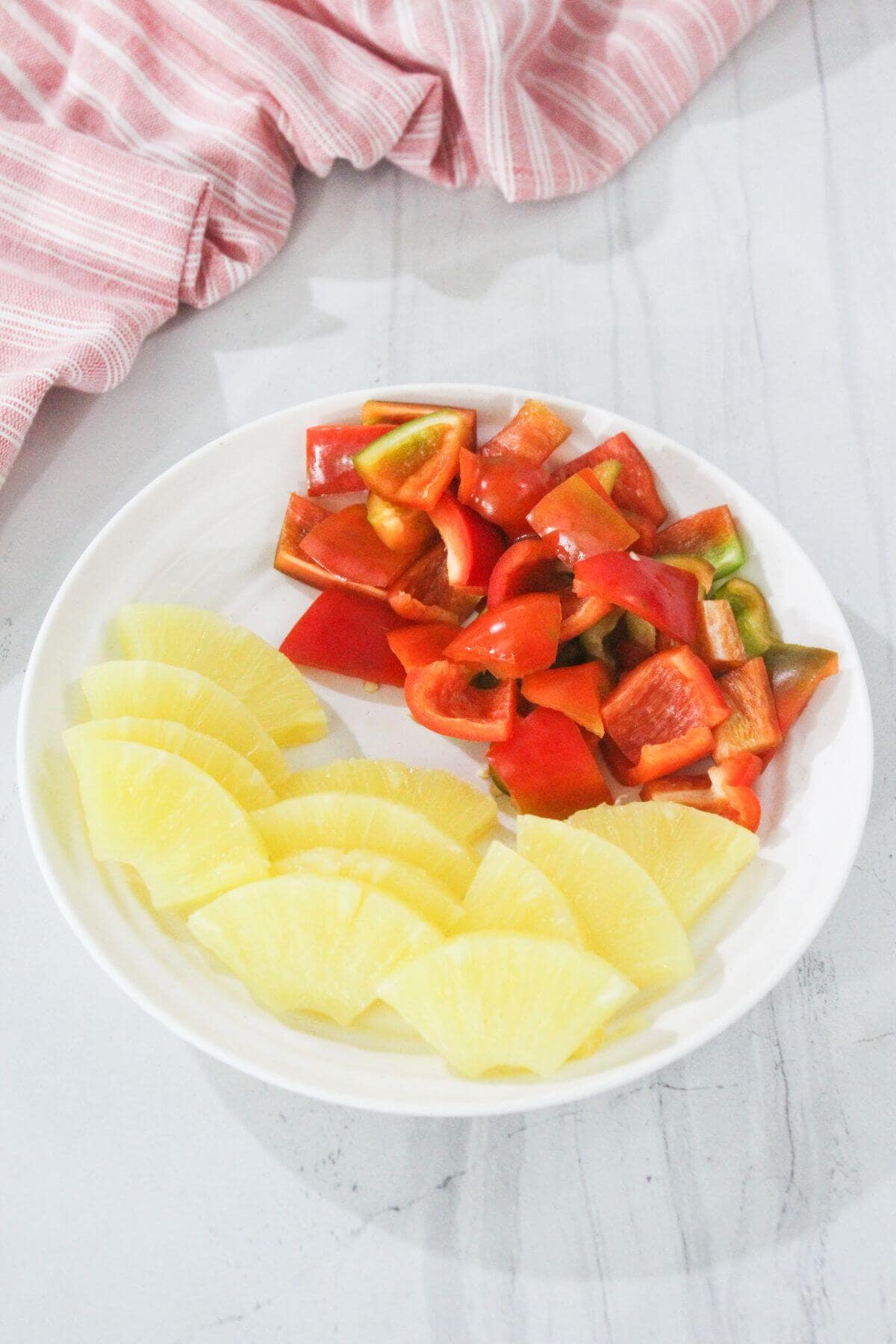 A white plate with sliced pineapple and chopped red and green bell peppers on a white marble surface with a pink striped cloth nearby.