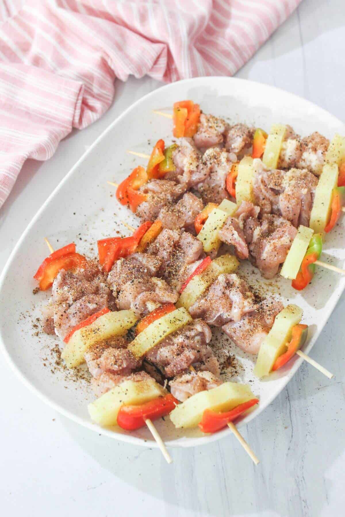 Raw chicken skewers with pineapple and bell peppers, seasoned with spices, arranged on a white oval plate; a pink-striped cloth in the background.