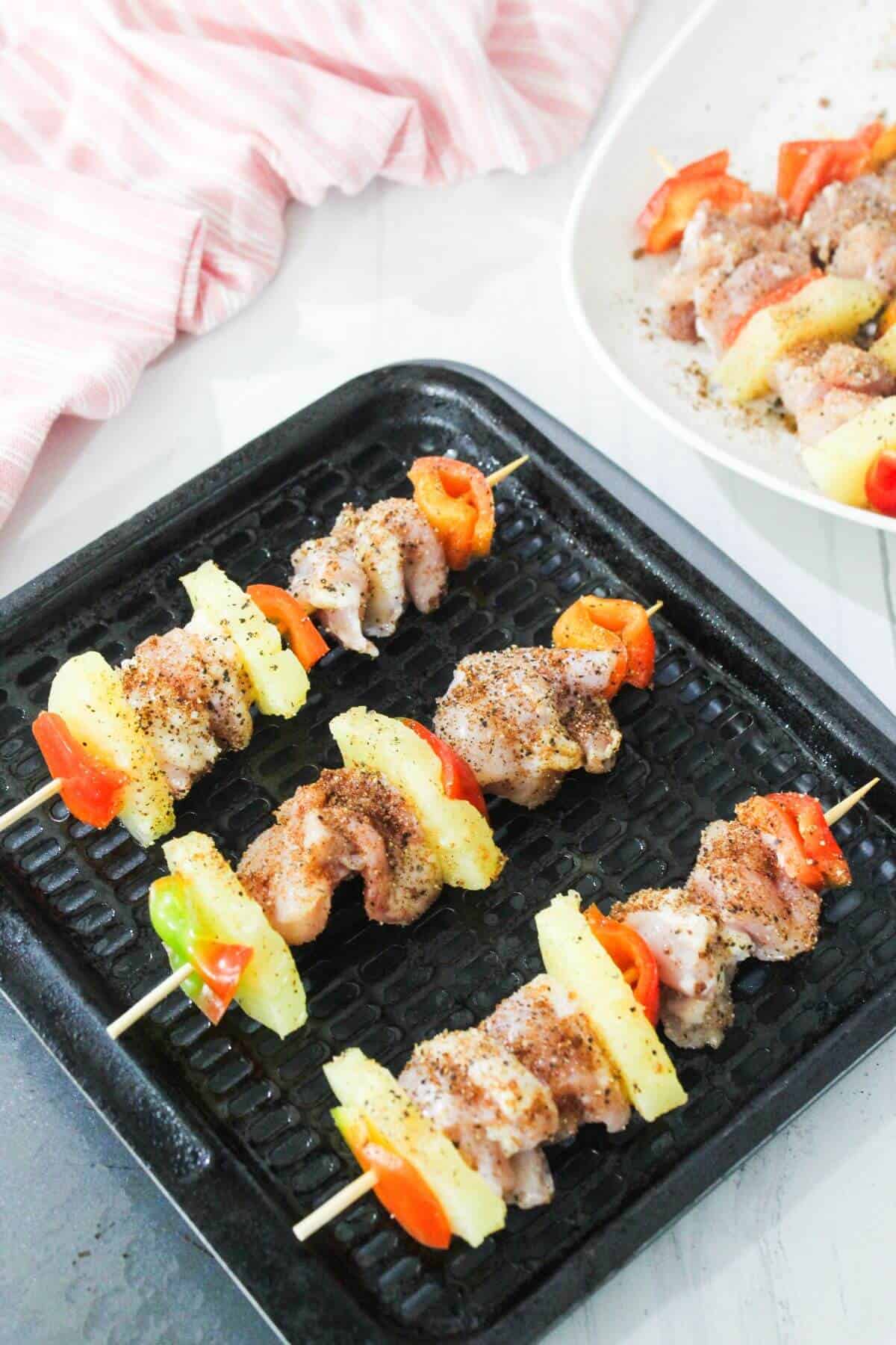 Skewers with pieces of raw chicken, pineapple, and bell pepper are arranged on a grill tray, ready to be cooked. A plate with more uncooked skewers is in the background.