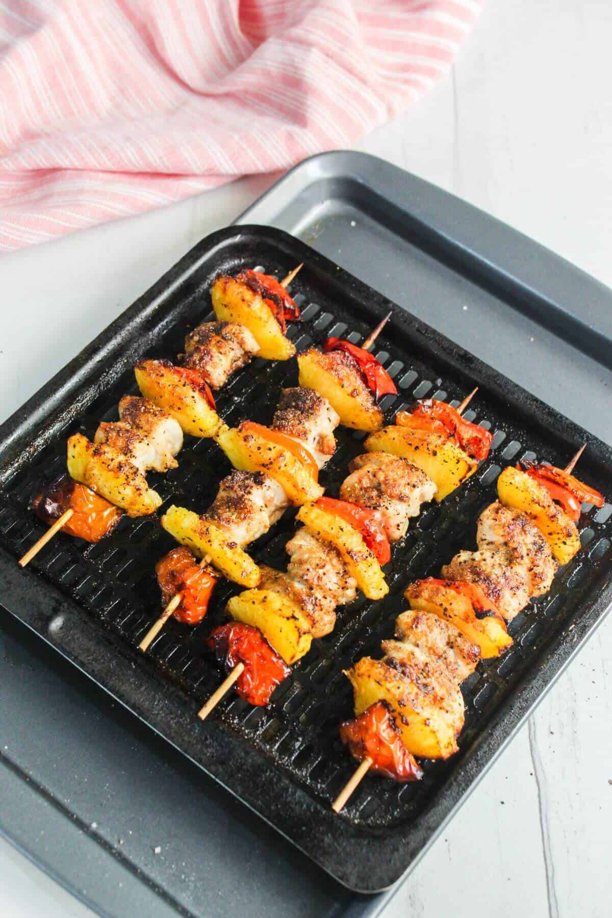 Three skewers with grilled chicken, yellow bell peppers, and tomatoes on a grill pan, with a pink-striped cloth in the background.