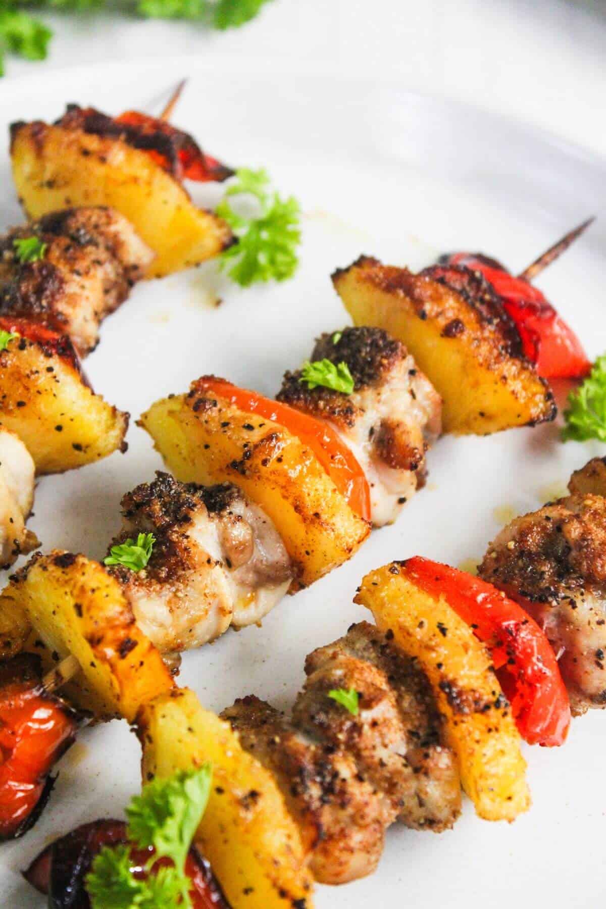 Grilled skewers with pieces of seasoned chicken, pineapple, and red bell pepper, garnished with fresh parsley on a white plate.