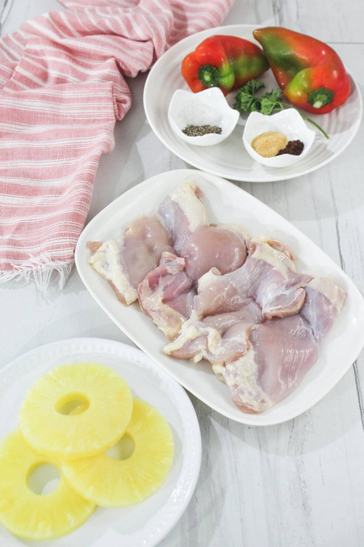 Raw chicken thighs on a plate, sliced pineapple rings, two whole bell peppers, small bowls of spices, and a pink striped cloth on a white surface.