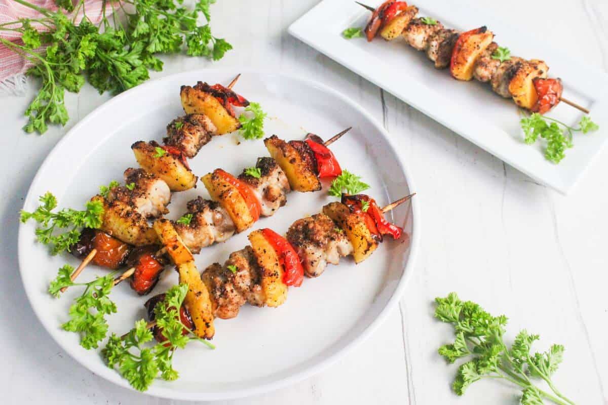 Three skewers with grilled chicken, bell peppers, and pineapple are served on a round white plate, garnished with parsley. Another skewer is on a rectangular plate nearby.