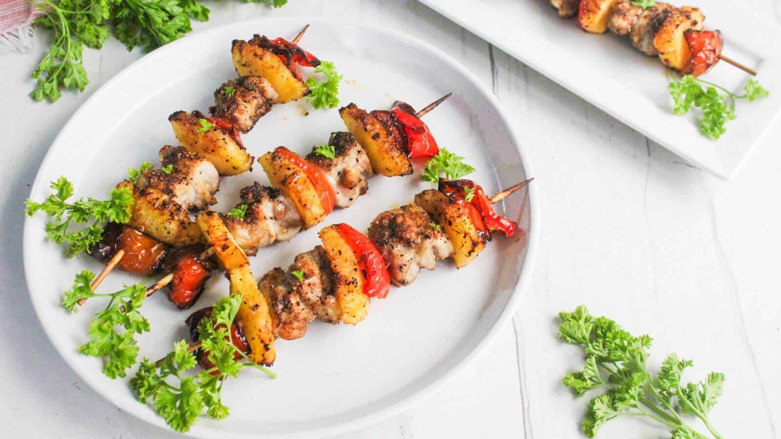 Three skewers with grilled meat, red bell peppers, and potatoes are served on a white plate, garnished with fresh parsley.