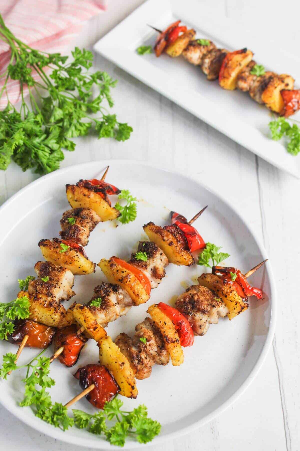 Three skewers with grilled chicken, pineapple, and red bell peppers are served on a white plate, garnished with parsley. Another skewer is on a rectangular platter in the background.