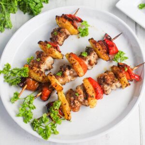 Two skewers with grilled chicken, pineapple, and red bell peppers are arranged on a white plate, garnished with fresh parsley.