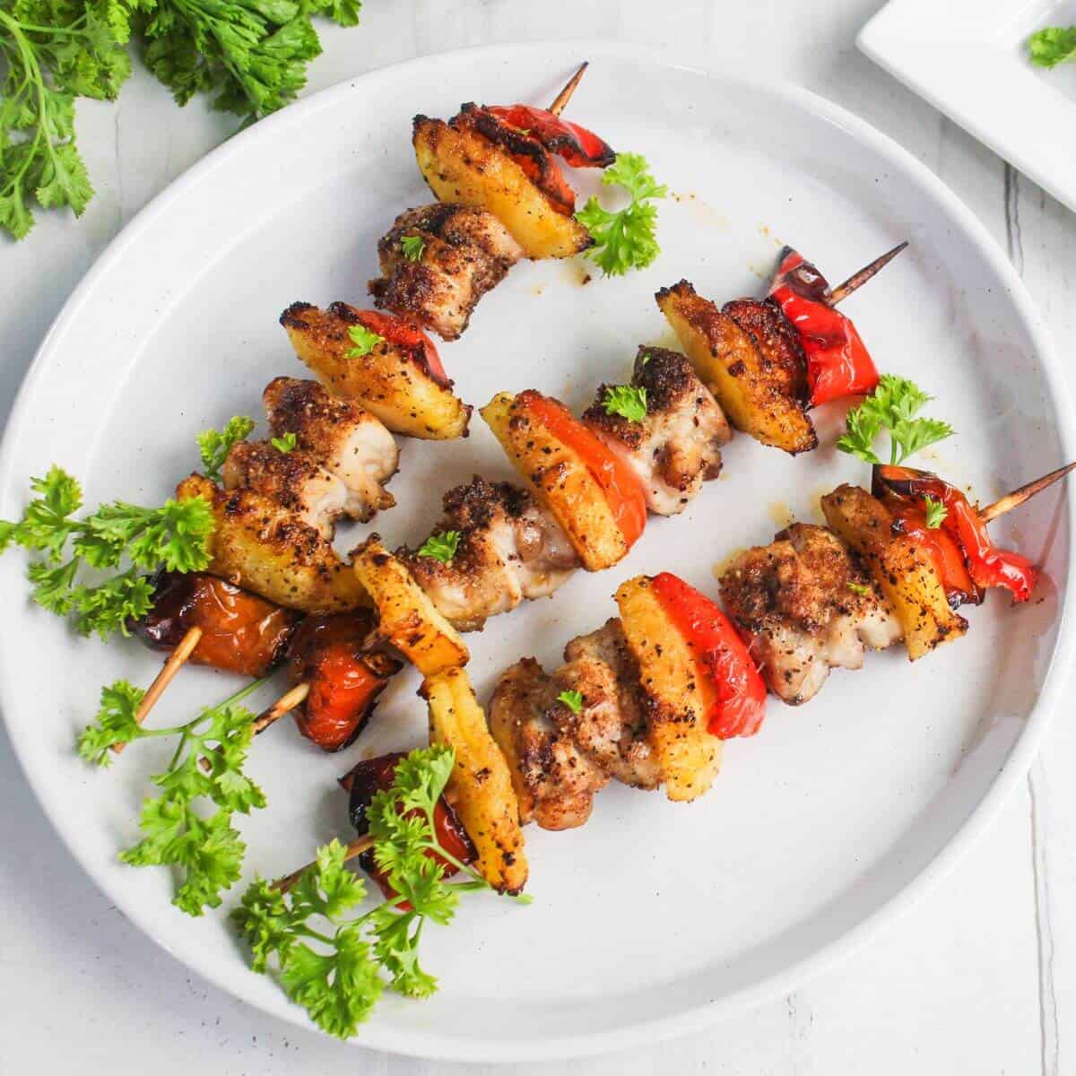 Two skewers with grilled chicken, pineapple, and red bell peppers are arranged on a white plate, garnished with fresh parsley.