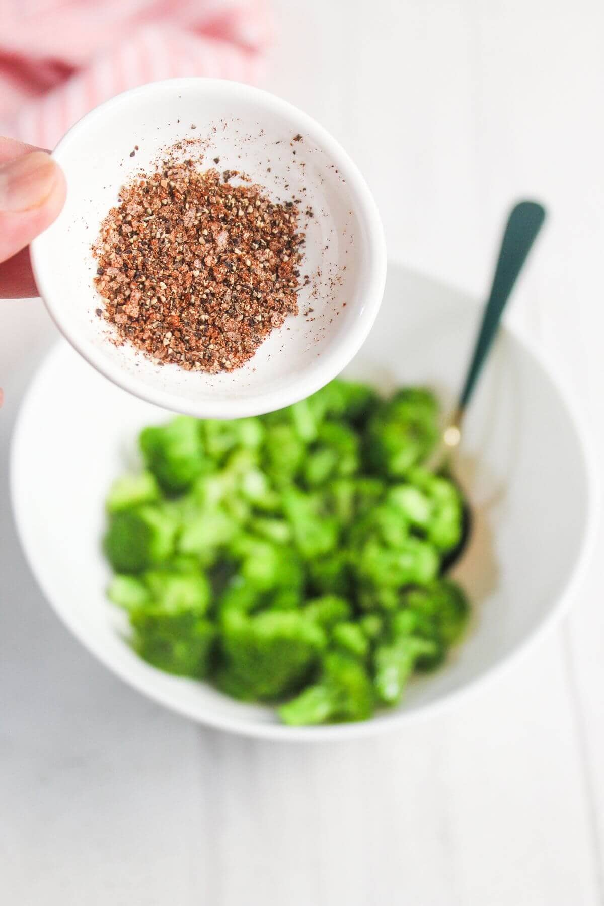 A hand holds a small white dish of seasoning above a bowl of fresh green lettuce with a fork inside.