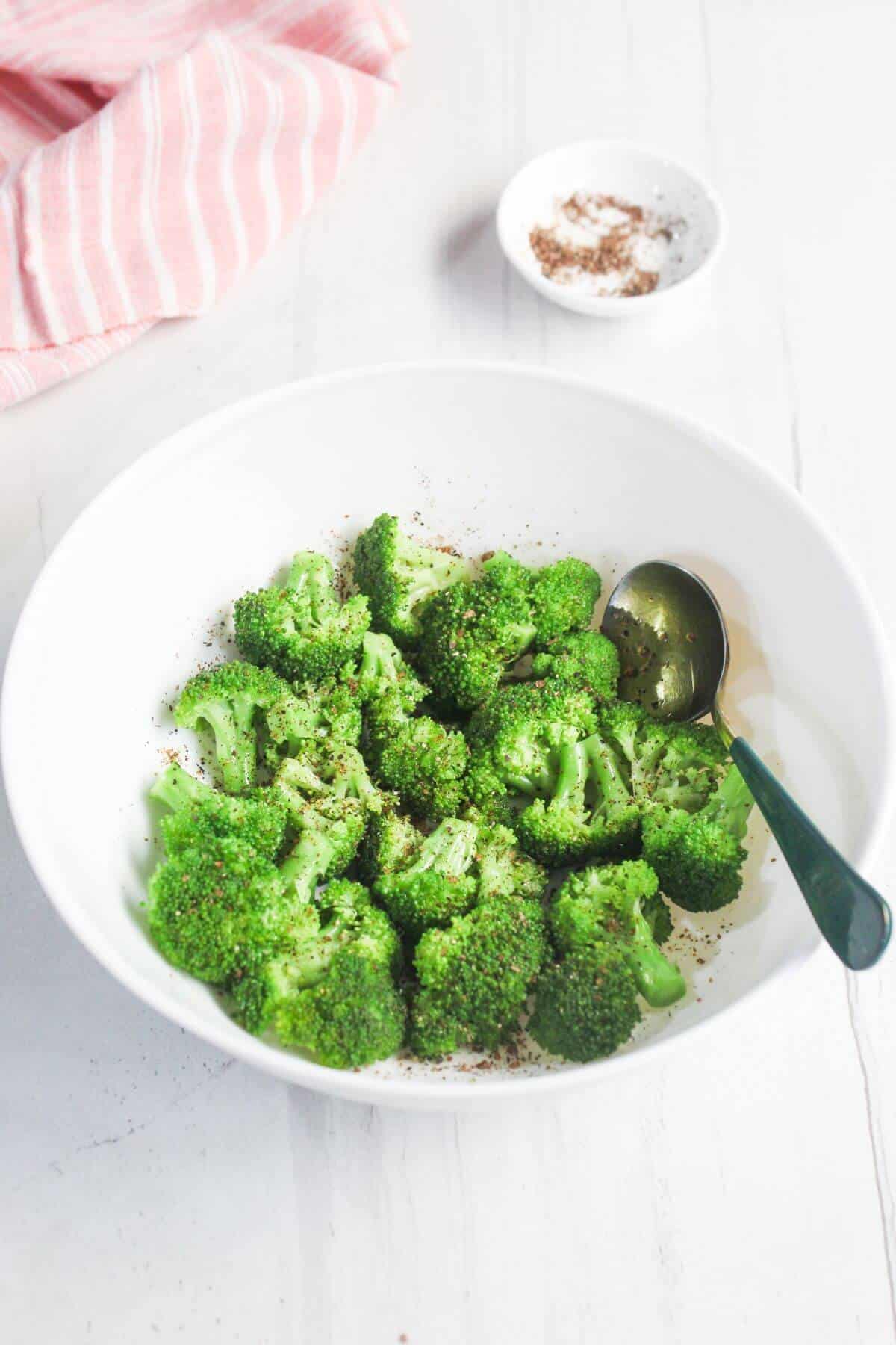 A white bowl filled with steamed broccoli florets, sprinkled with black pepper, with a spoon inside; a small dish of seasoning and a striped cloth are in the background.