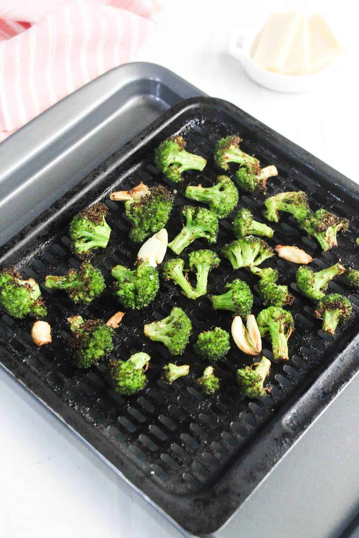Roasted broccoli florets and garlic cloves on a black baking tray, with a pink-striped towel and a small white dish in the background.