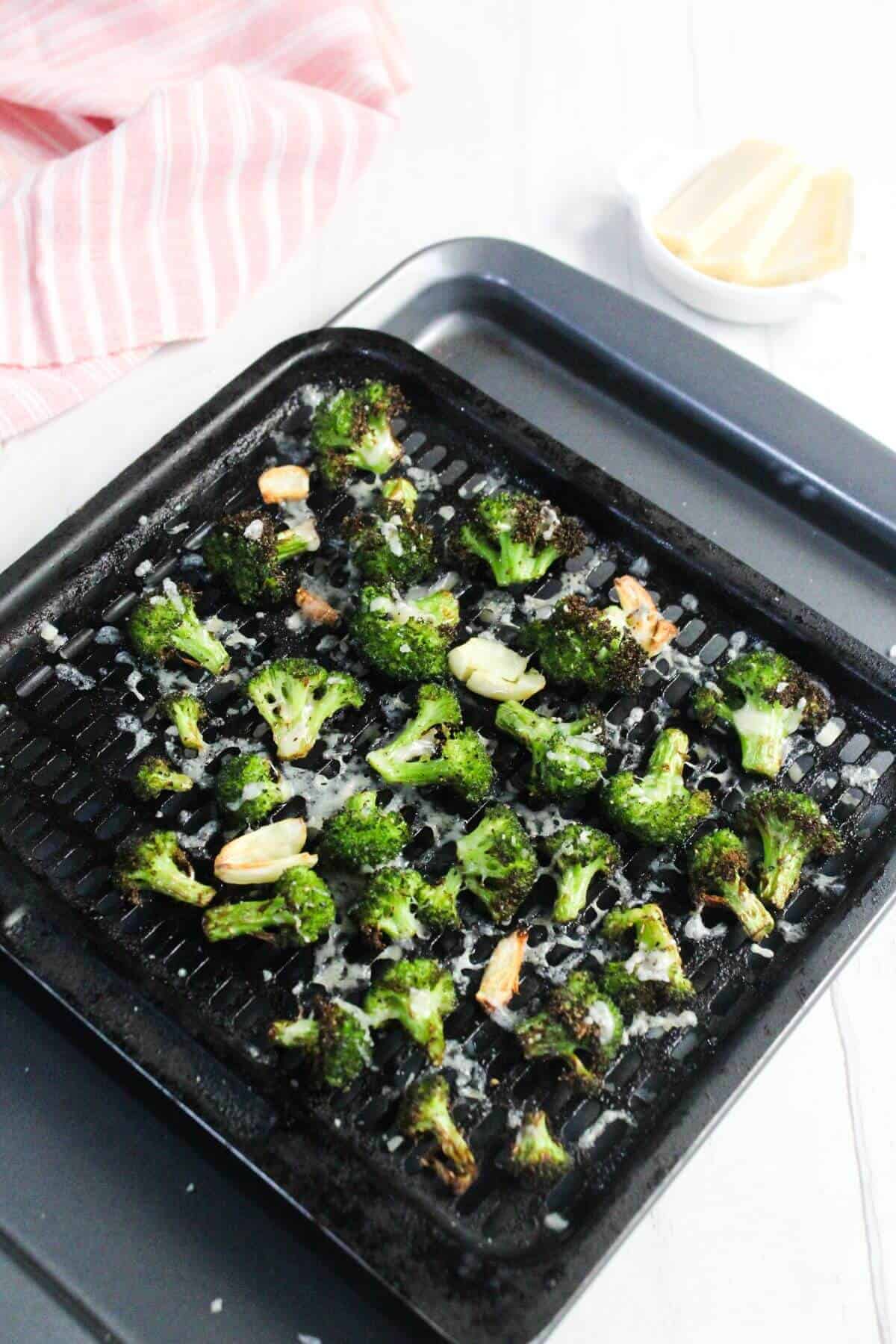 Roasted broccoli florets and garlic cloves on a black grill pan, with shredded cheese sprinkled on top. A pink striped towel and a dish of cheese are in the background.