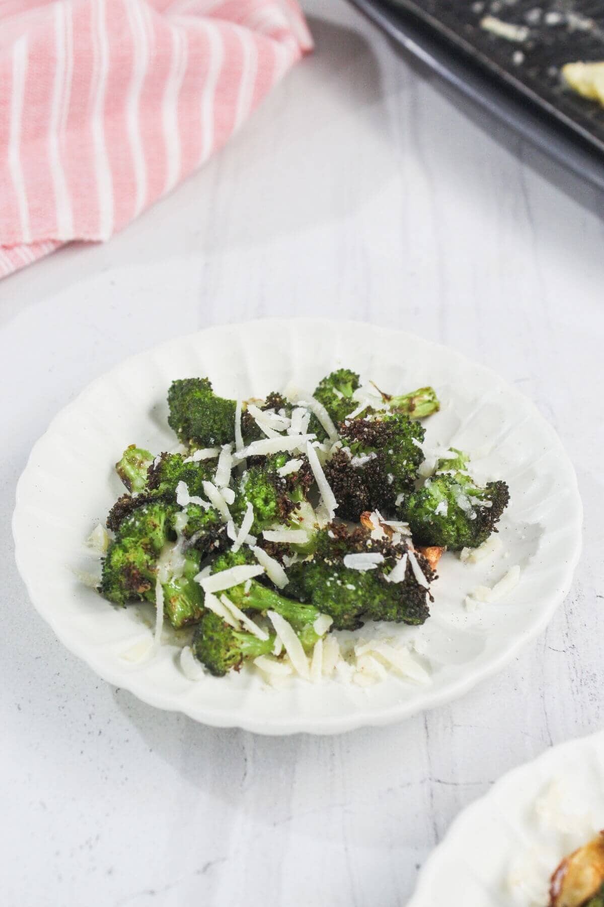 Roasted broccoli florets topped with shredded cheese on a white plate, placed on a light surface near a pink-striped cloth.