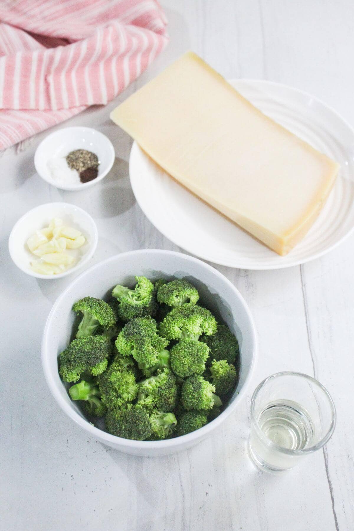 A bowl of broccoli florets, a glass of clear liquid, sliced garlic, a plate with a block of cheese, and a small bowl of salt and pepper on a white surface.