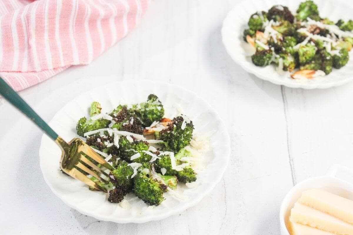 A fork holds roasted broccoli topped with grated cheese on a white plate; another plate and a bowl with cheese are nearby on a white surface.
