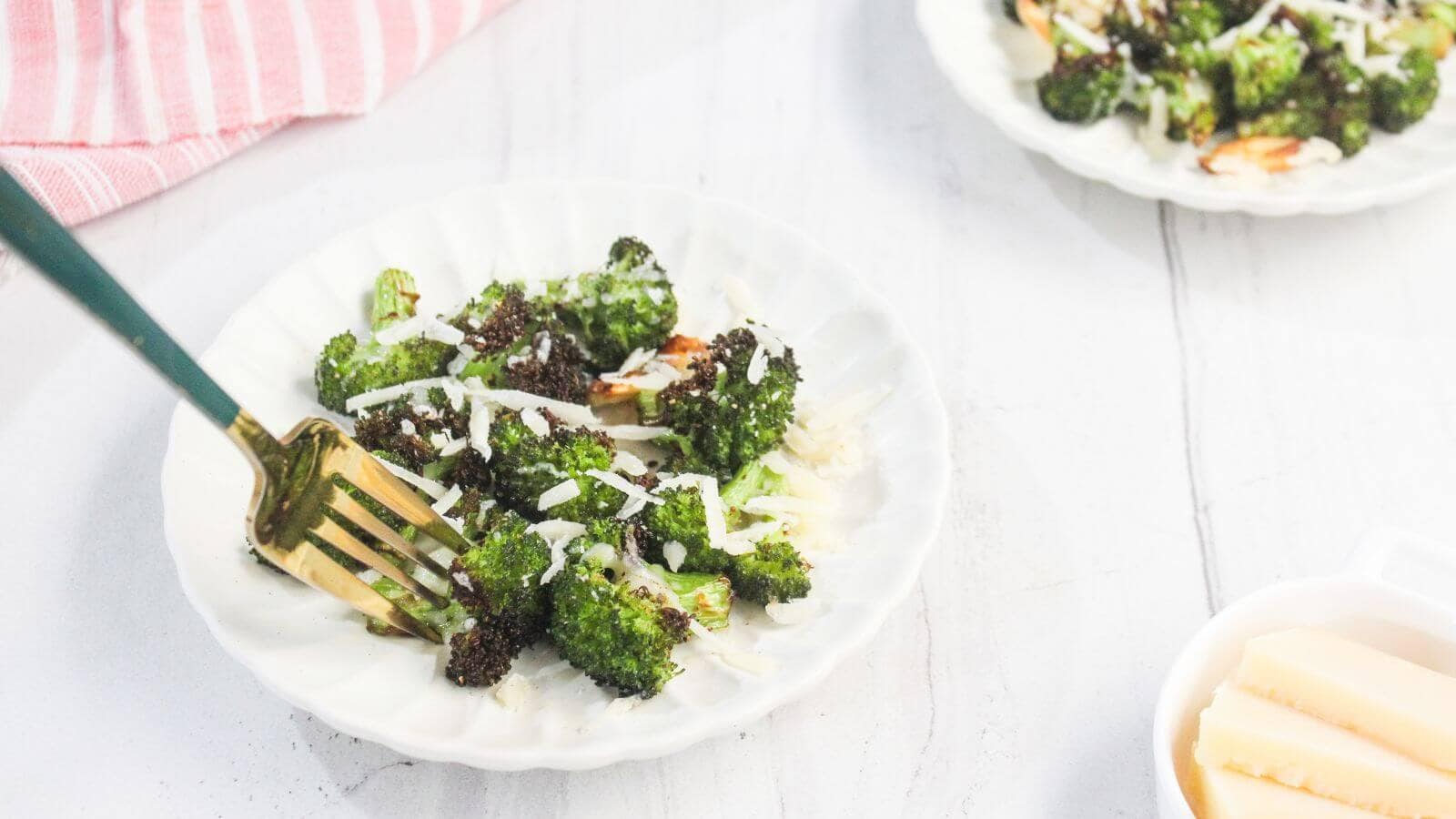A fork rests on a white plate with roasted broccoli topped with grated cheese; another plate of broccoli and a bowl with cheese are nearby.
