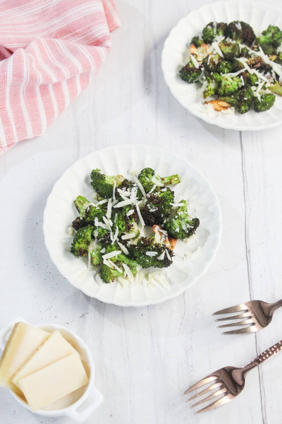 Two white plates with roasted broccoli topped with shredded cheese on a white surface, with a dish of sliced cheese, two forks, and a pink striped napkin beside them.