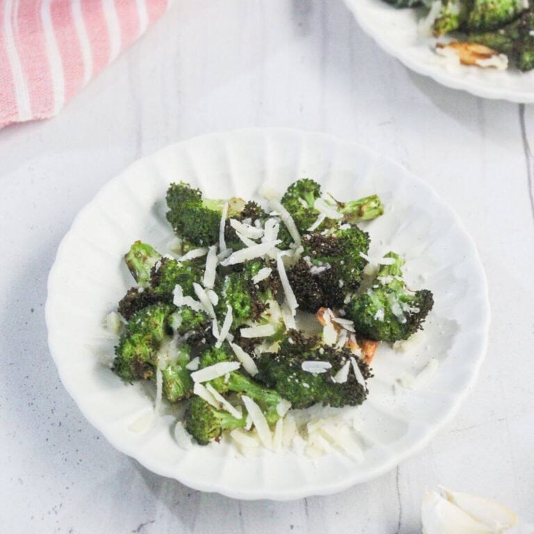A white plate with roasted broccoli florets topped with shredded cheese, placed on a white surface next to a pink-striped cloth.