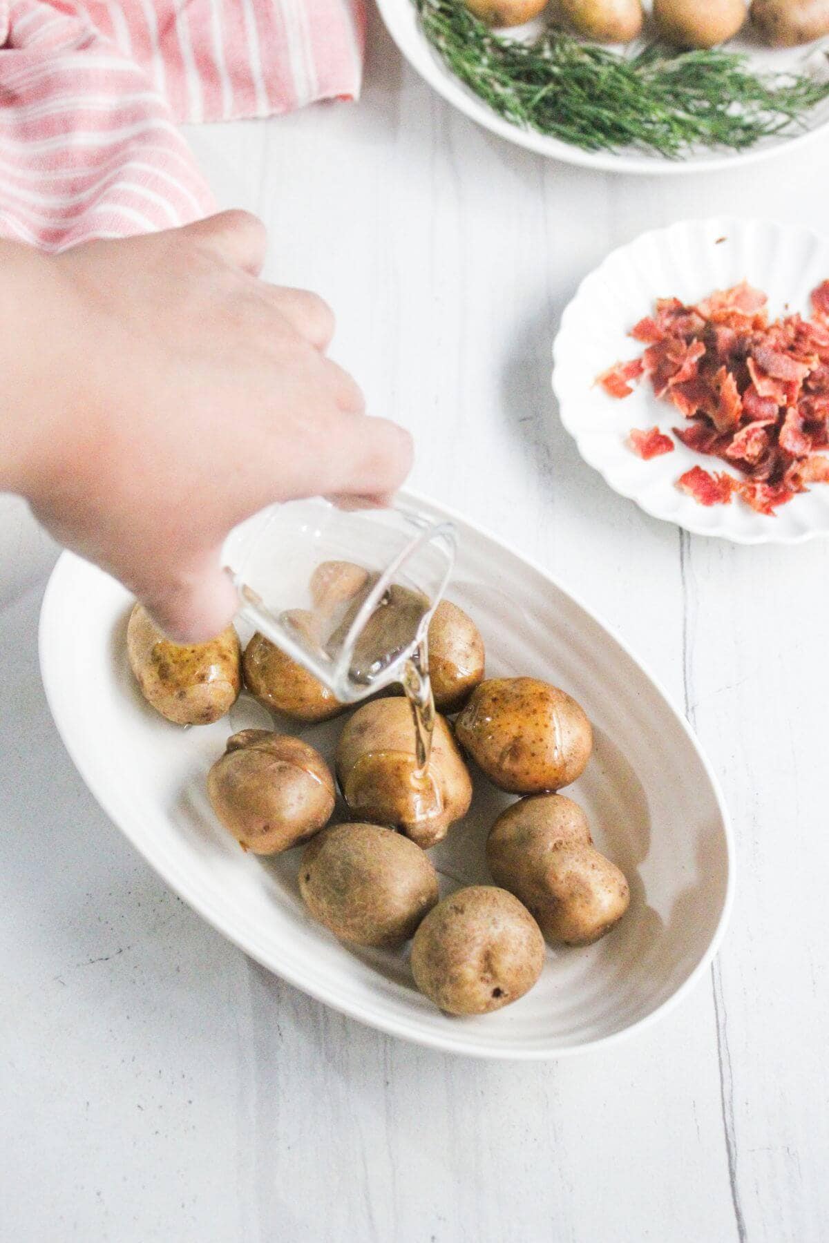 A hand pours liquid from a small glass pitcher over whole baby potatoes in a white oval dish, with dried herbs and chopped bacon in the background.