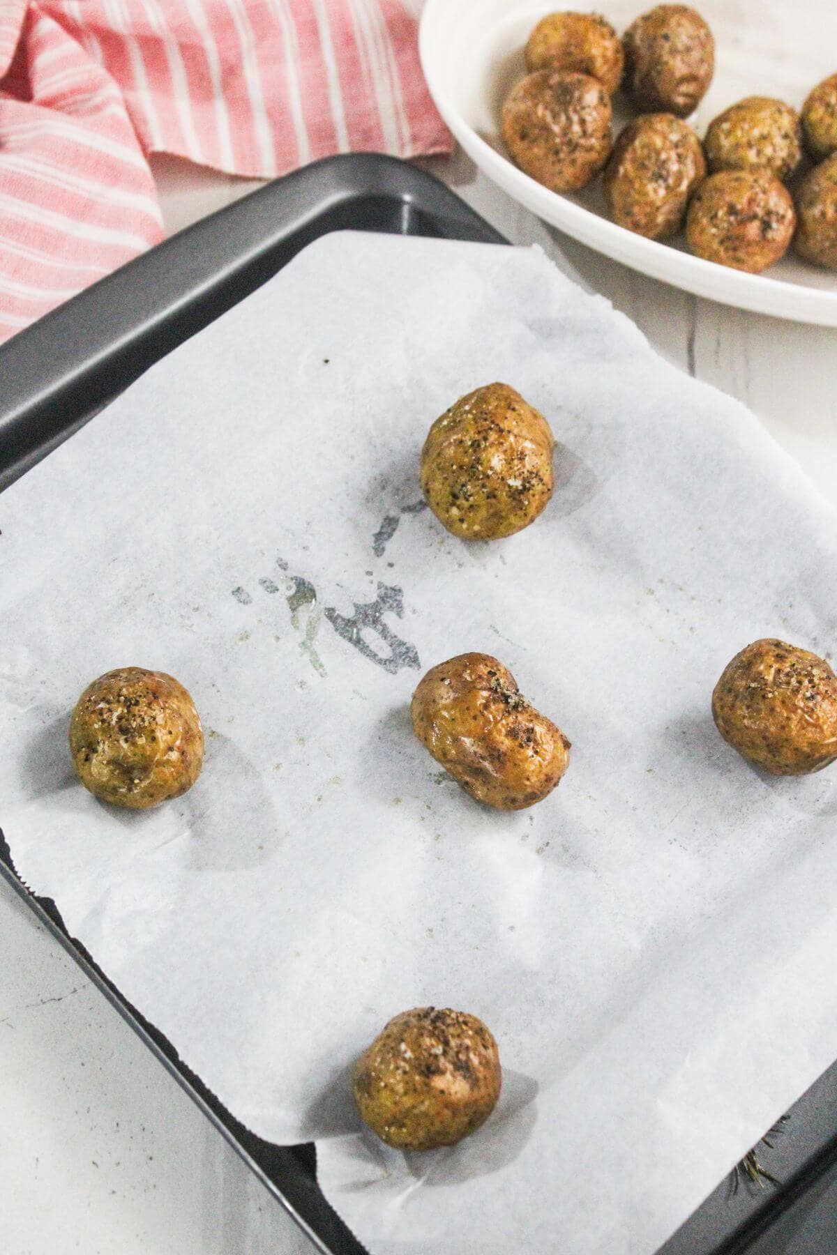 Six baked meatballs are arranged on a parchment-lined baking tray, with more meatballs visible in a white dish in the background. A pink striped cloth is partially visible.