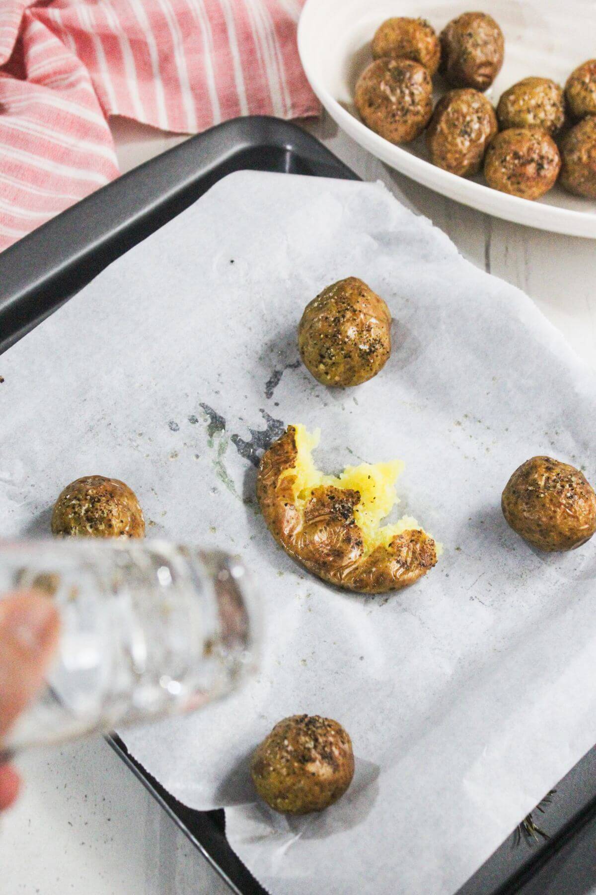 A hand sprinkles salt onto smashed roasted baby potatoes on a parchment-lined baking tray, with a bowl of whole roasted potatoes in the background.