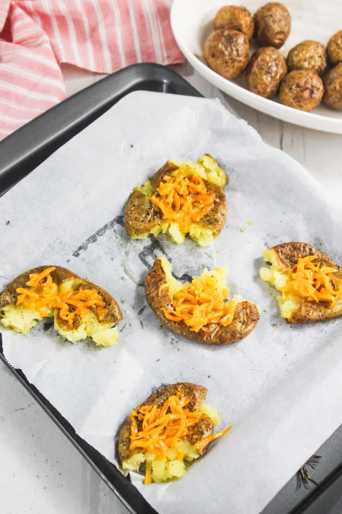 A baking tray lined with parchment paper holds five smashed potatoes topped with shredded cheese; a dish of whole roasted potatoes is in the background.