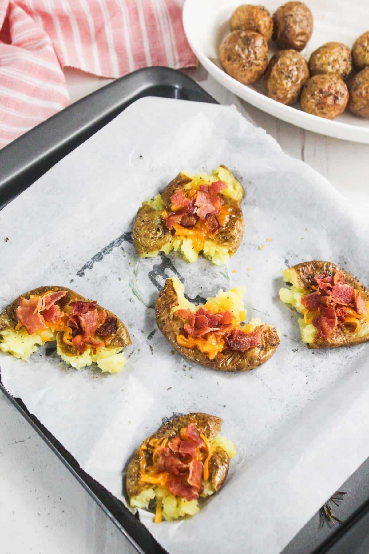Five smashed potatoes with melted cheese and bacon bits are on a parchment-lined baking tray; a dish of whole roasted potatoes is in the background.